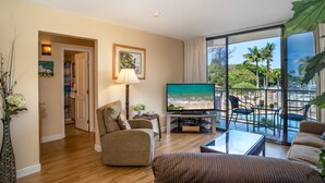 A living room with a TV, a sofa, a coffee table, and a balcony with a view of palm trees.
