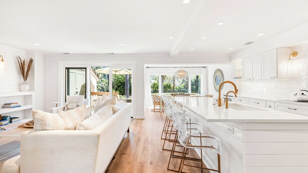 Una cocina moderna con armarios blancos, una isla grande con taburetes y vistas al exterior gracias a amplias ventanas.