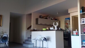 A living room with a wooden coffee table, a white chair, and a kitchen area with a counter and cabinets. - Duplex 160 m2 (Saint Gilles les bains)