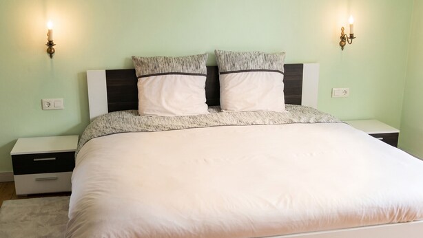 A neatly made bed with a white comforter and two pillows, a bedside table with a lamp, and a wall-mounted light fixture. - Homerez - House in Berrwiller (Berrwiller)