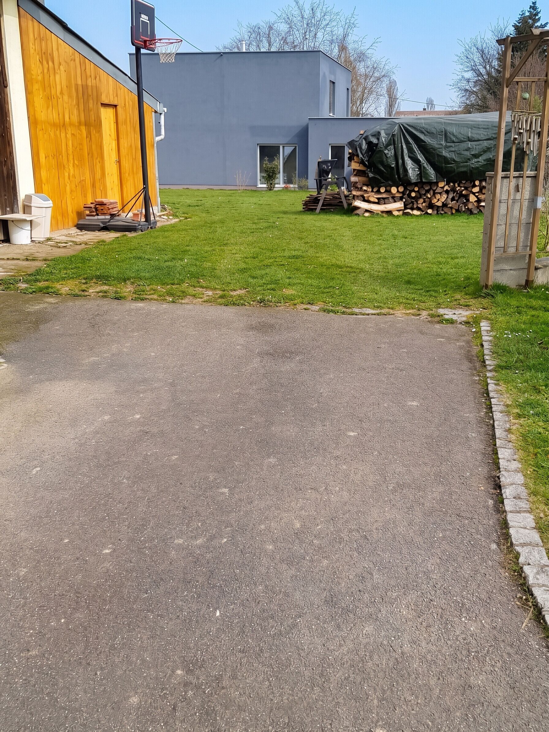 A paved driveway leading to a building with a basketball hoop, a wooden fence, and a stack of firewood.