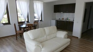 A living room with a white sofa, wooden dining table, and chairs. There is a kitchen area with dark cabinets and a tiled backsplash. - Inviting 1-bed Apartment in Aleksandrovo (Pomorie)