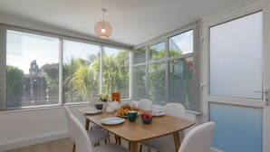 Un coin repas avec une table en bois, des chaises blanches et une vue sur la verdure grâce à de grandes fenêtres.