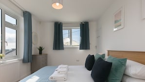 Une chambre à coucher avec un lit, une fenêtre, des rideaux, une table de chevet avec une plante et un tableau accroché au mur.