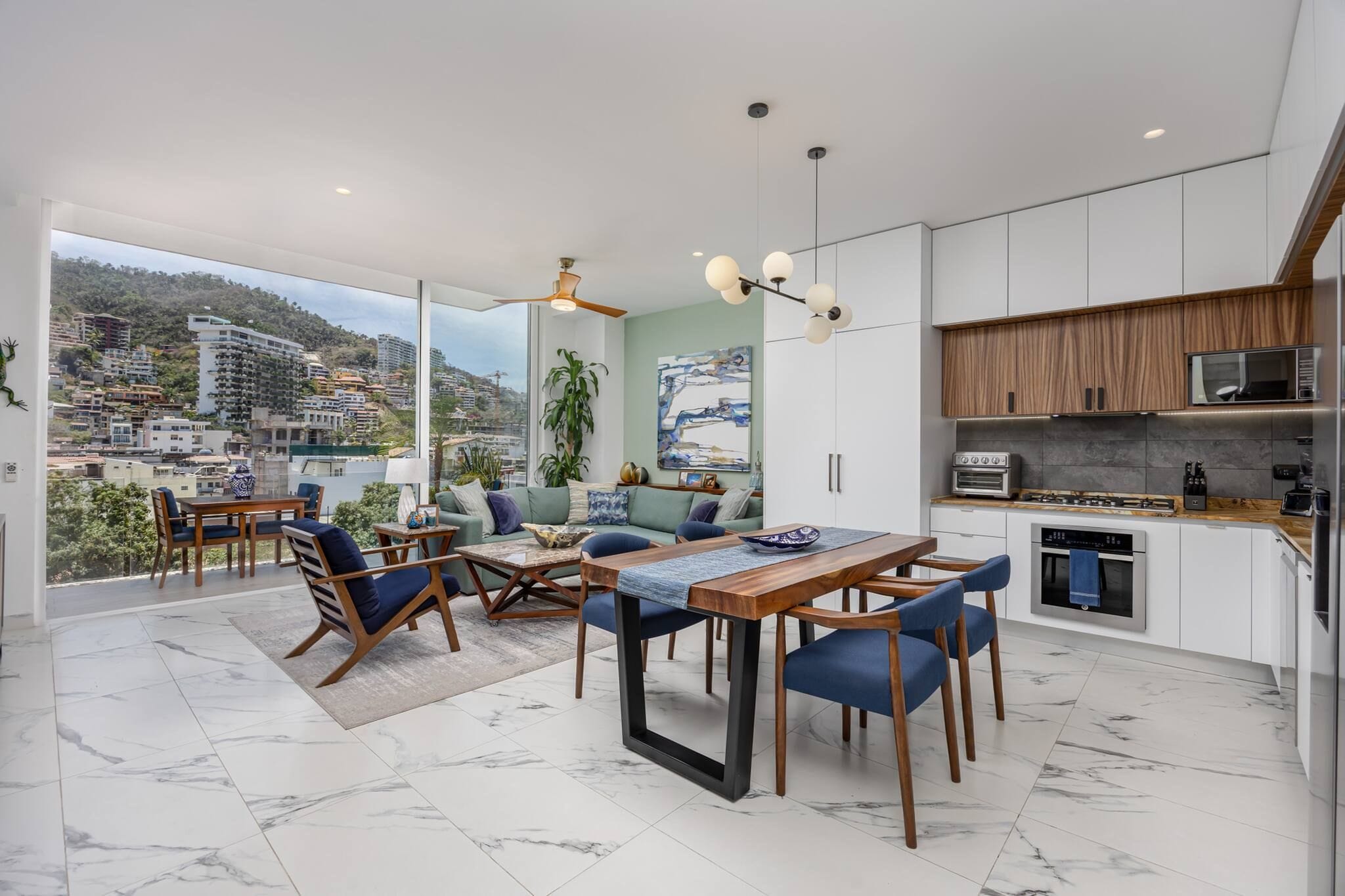 A modern kitchen with white cabinets and a wooden countertop, a dining area with a table and chairs, and a balcony with a view of a hillside town.