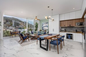 A modern kitchen with white cabinets and a wooden countertop, a dining area with a table and chairs, and a balcony with a view of a hillside town.