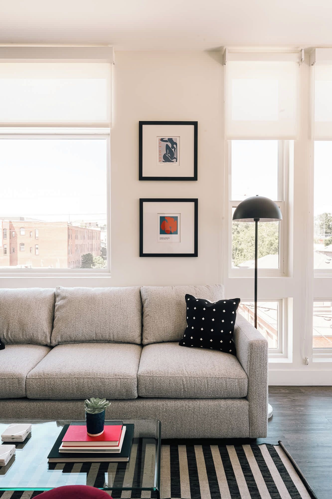Sala de estar con un sofá color beige, dos cuadros enmarcados, una lámpara de pie negra y vistas de edificios a través de grandes ventanales.