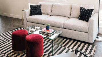 Una sala de estar con un sofá color beige, alfombra estampada en blanco y negro, otomanas de terciopelo rojo y una mesa de centro de vidrio con libros y una planta.