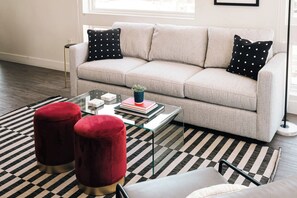 A living room with a beige sofa, black and white patterned rug, red velvet ottomans, and a glass coffee table with books and a plant.
