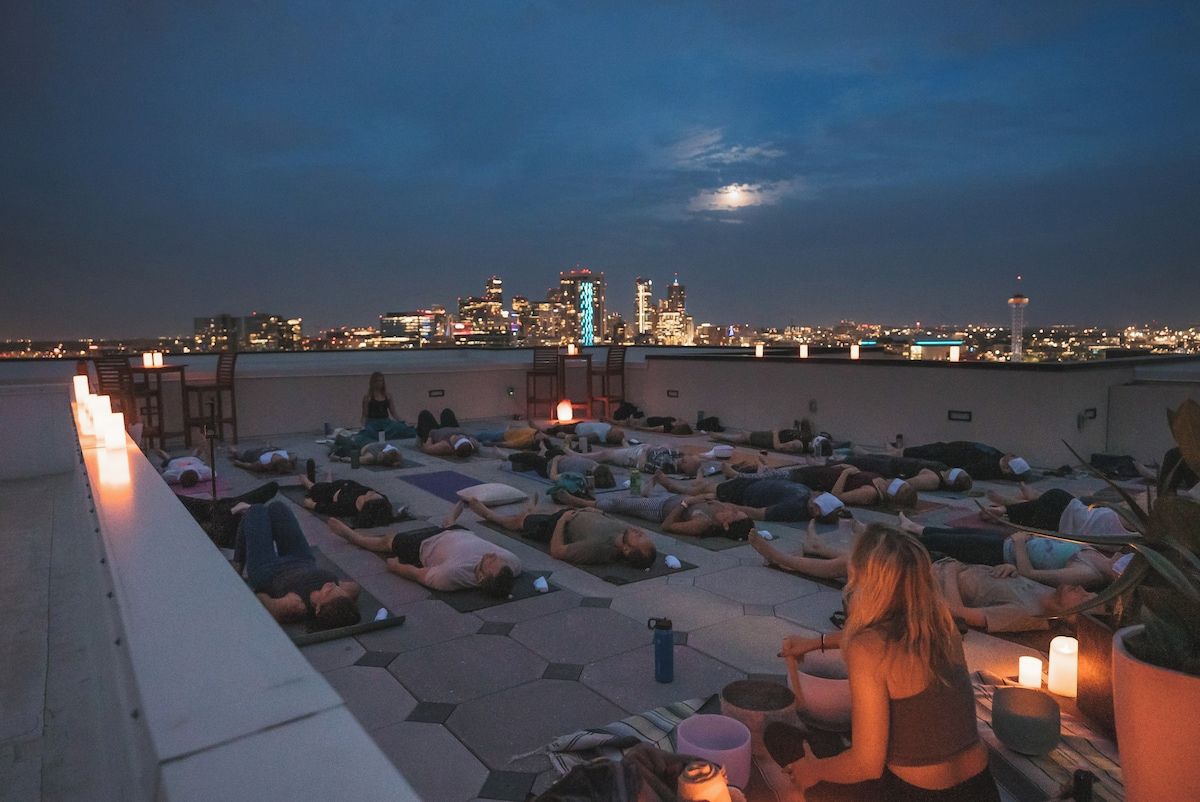 Eine Dachterrassenfeier mit Menschen, die auf Matten liegen, einige lesen, und einer Stadtansicht im Hintergrund.