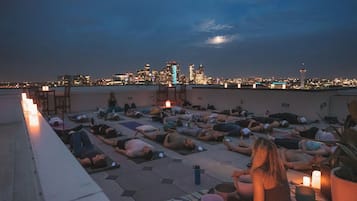 Eine Dachterrassenfeier mit Menschen, die auf Matten liegen, einige lesen, und einer Stadtansicht im Hintergrund.