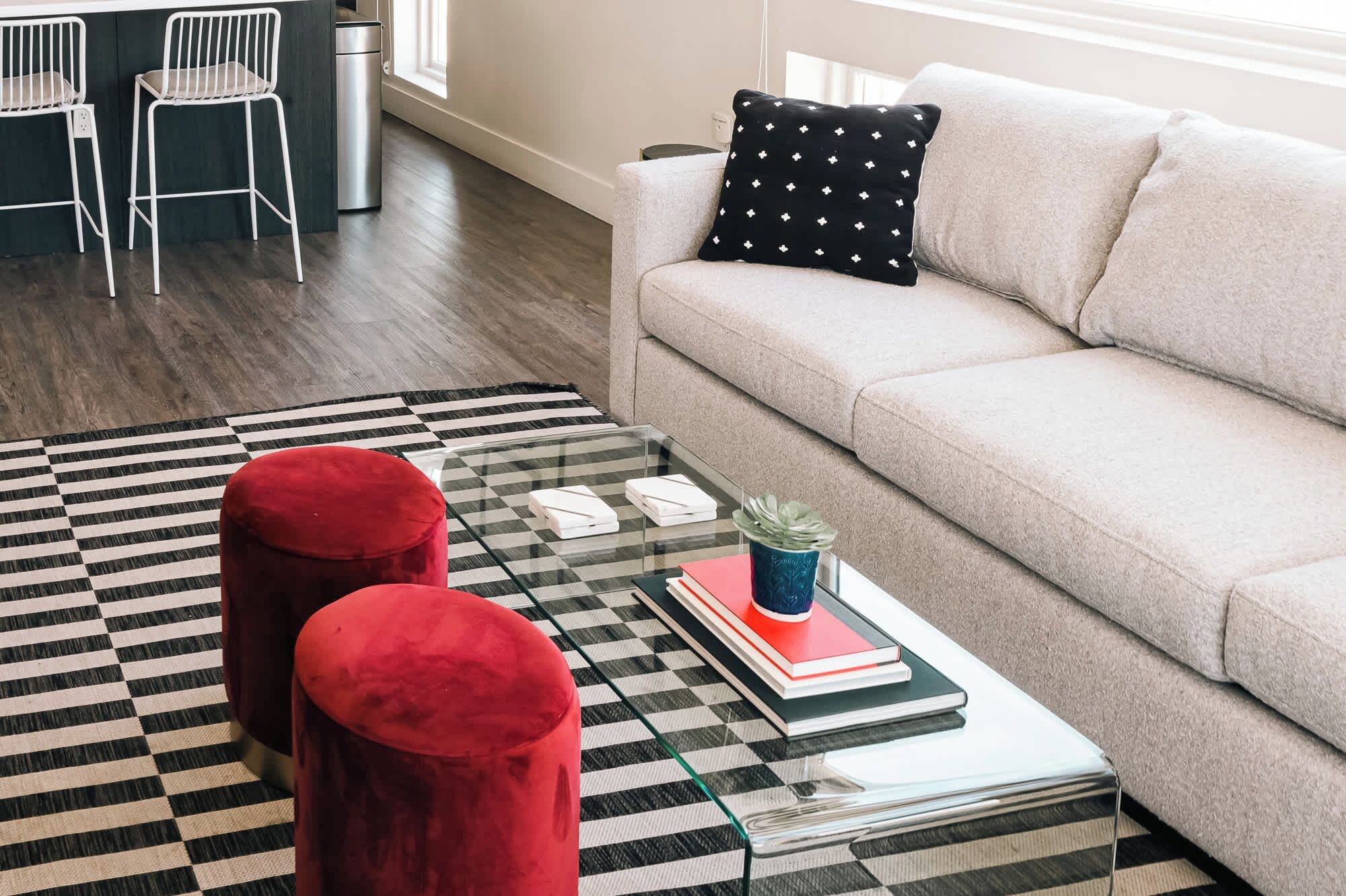 A living room with a beige sofa, a black and white checkered rug, two red ottomans, a glass coffee table with books and a plant, and a black and white pillow.