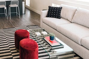 A living room with a beige sofa, a black and white checkered rug, two red ottomans, a glass coffee table with books and a plant, and a black and white pillow.