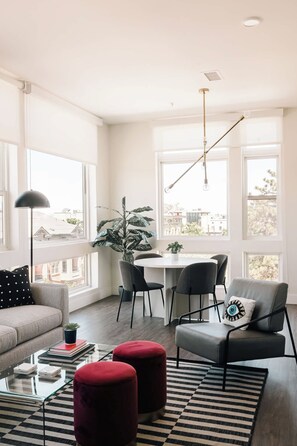 A modern living room with a sofa, a dining table with chairs, a coffee table, and a large window with a view of buildings. - A Luxe Point of View | Pet-Friendly | Zuni Lofts (Denver)