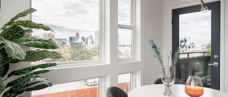 A room with large windows, a round table, a potted plant, and a vase with flowers.