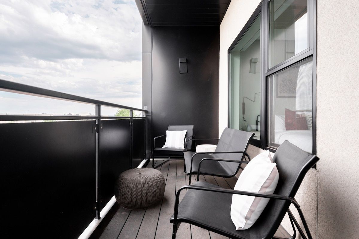 Ein Balkon mit zwei schwarzen Stühlen, einem runden Hocker, einer Glastür und Blick zum Himmel.
