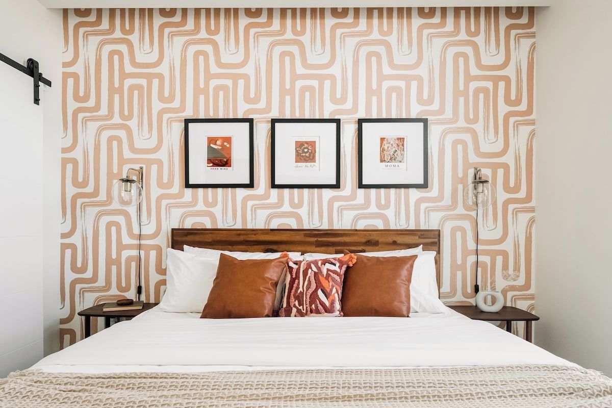 Una cama bien hecha con un papel tapiz estampado en la pared de fondo, una mesita de noche con una lámpara, y tres cuadros enmarcados en la pared.