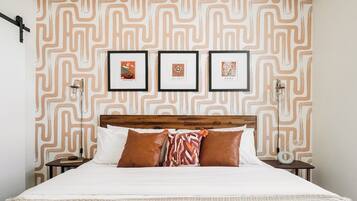 Una cama bien hecha con un papel tapiz estampado en la pared de fondo, una mesita de noche con una lámpara, y tres cuadros enmarcados en la pared.