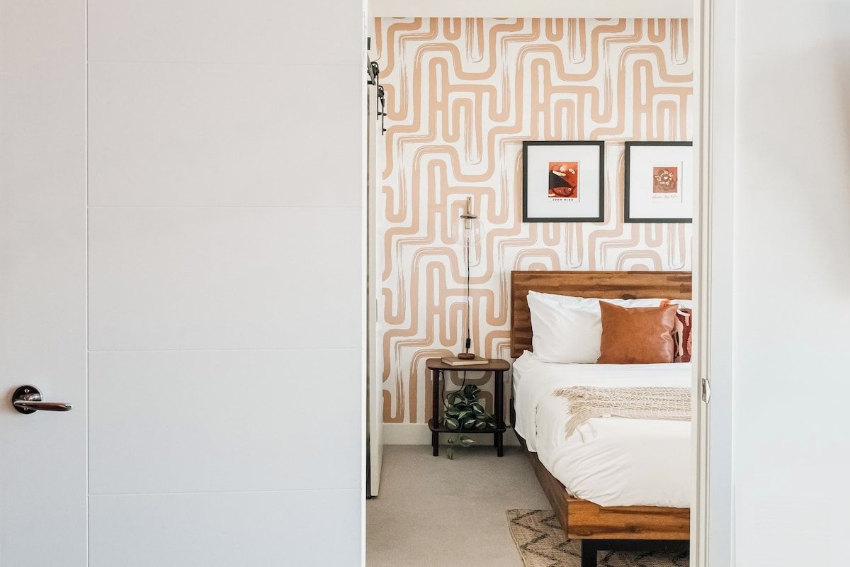 A bedroom with a patterned wallpaper, a bed with white bedding, a nightstand with a lamp, and two framed pictures on the wall.