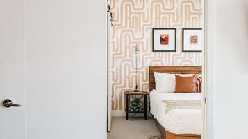 A bedroom with a patterned wallpaper, a bed with white bedding, a nightstand with a lamp, and two framed pictures on the wall.