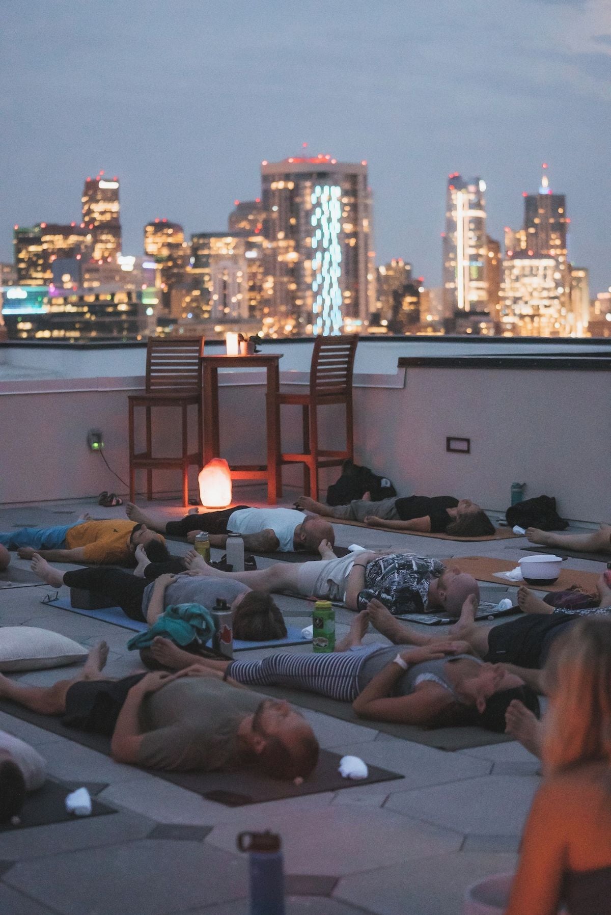 A rooftop relaxation area with people lying on mats, a table with a lamp, and a city skyline in the background.
