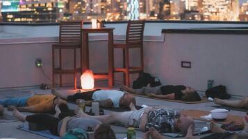 A rooftop relaxation area with people lying on mats, a table with a lamp, and a city skyline in the background.