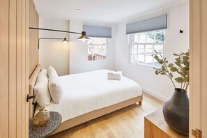 Ein modernes Schlafzimmer mit Bett, Nachttischlampen, einem Fenster mit Jalousien und einer Zimmerpflanze.