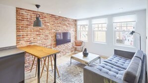 A modern living room with a brick wall, a wooden table, a grey sofa, and a TV mounted on the wall. - Host & Stay | Modern Industrial (Whitby)