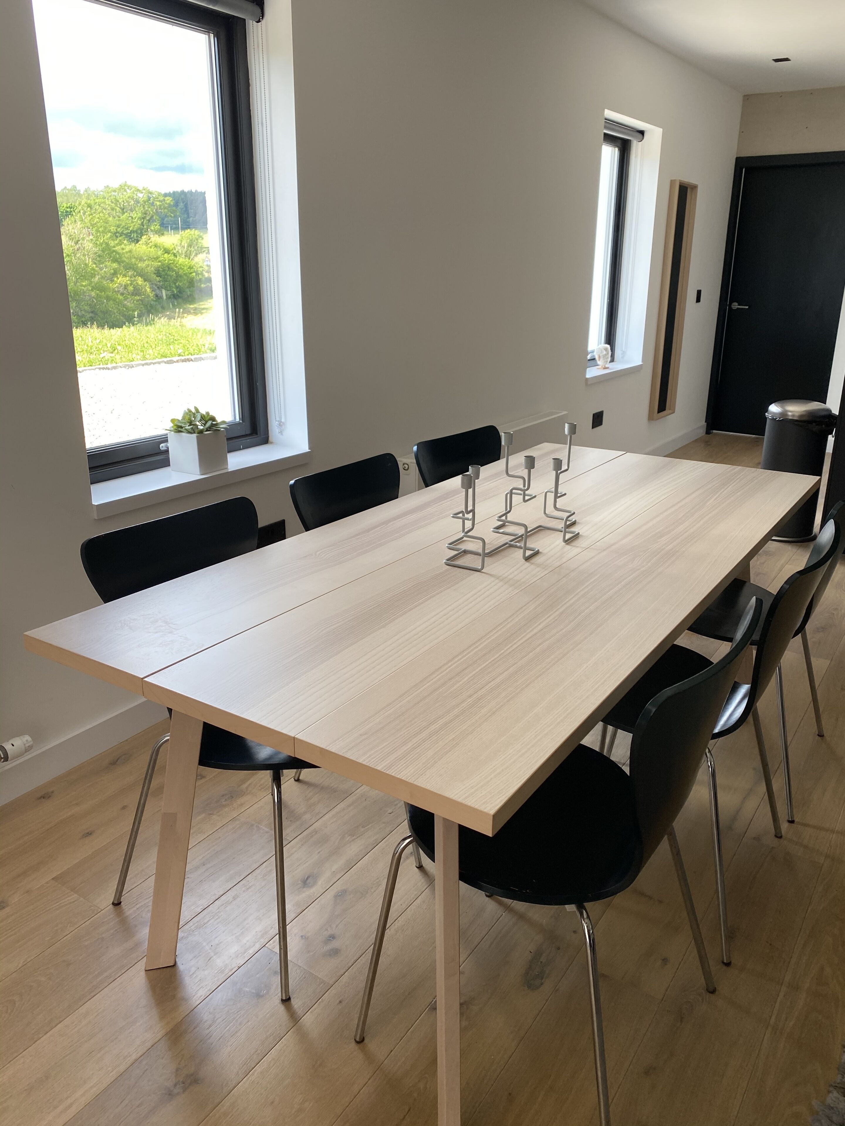 A modern dining area with a light wood table, black chairs, and a window with a view of greenery.