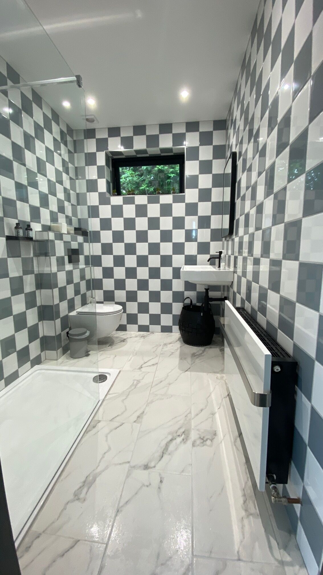 A modern bathroom with a checkered tile wall, a toilet, a sink, a shower, and a window with a view of greenery.