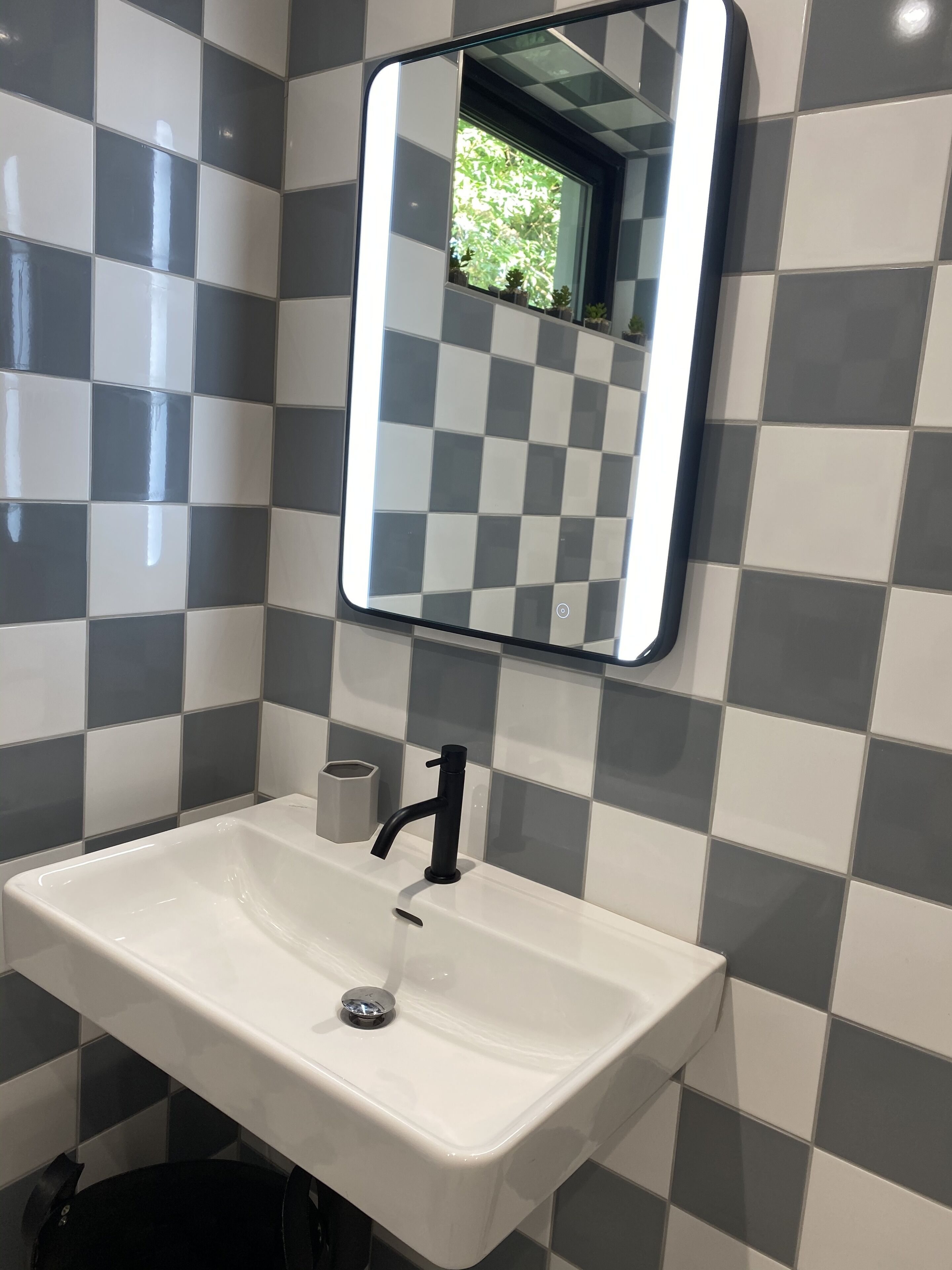 Un baño con un lavamanos blanco, grifo negro y un espejo encima. Las paredes están cubiertas con un estampado de cuadros.