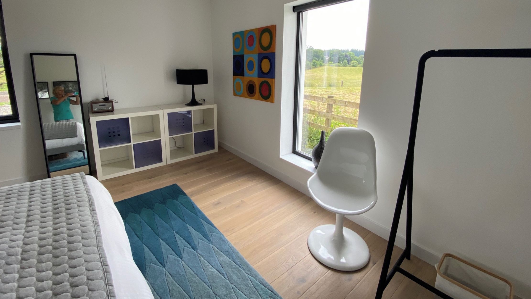 A bedroom with a bed, a desk, a chair, a window with a view of the outdoors, and a wall with colorful artwork.