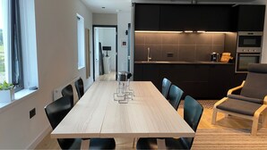 Ett modernt matrum med ett långt träbord och svarta stolar, ett kök med mörka skåp och en inbyggd ugn, och en utsikt över naturen genom ett fönster.
