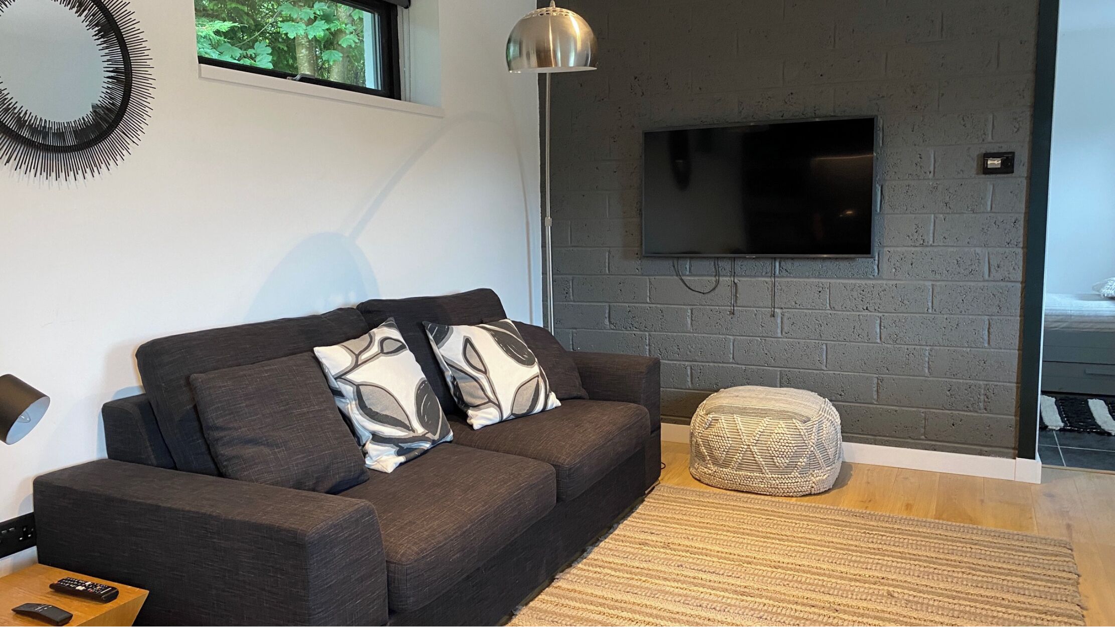 A modern living room with a dark grey sofa, patterned cushions, a round woven ottoman, a floor lamp, and a flat-screen TV mounted on a grey brick wall.
