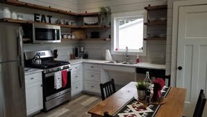 Eine moderne Küche mit weißen Schränken, einem Edelstahlkühlschrank, einer Mikrowelle, einem Herd und einem Spülbecken. Darüber befinden sich Holzregale und ein Fenster sorgt für natürliches Licht.