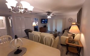 A living room with a dining area, a television, and a ceiling fan. - Sand Pebbles #1401 Chateau La Mer II 2Bd-2Bath Walk-In Shower Ground Floor (Destin)