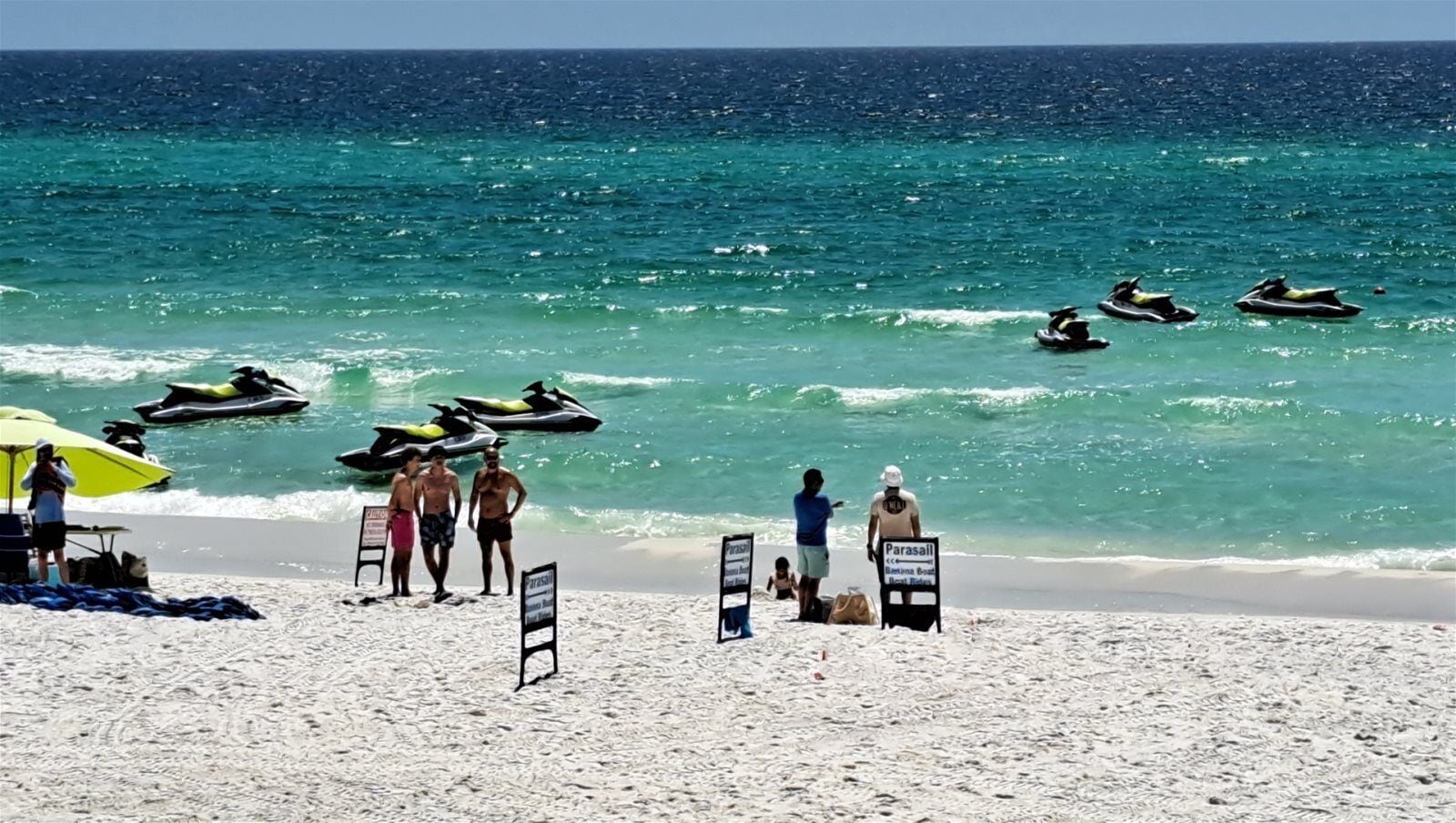 A beach with jet skis, a parasail sign, and people on the sand.