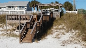 A wooden staircase leading to a beach house with a white fence and palm trees in the background. - Sand Pebbles #1401 Chateau La Mer II 2Bd-2Bath Walk-In Shower Ground Floor (Destin)