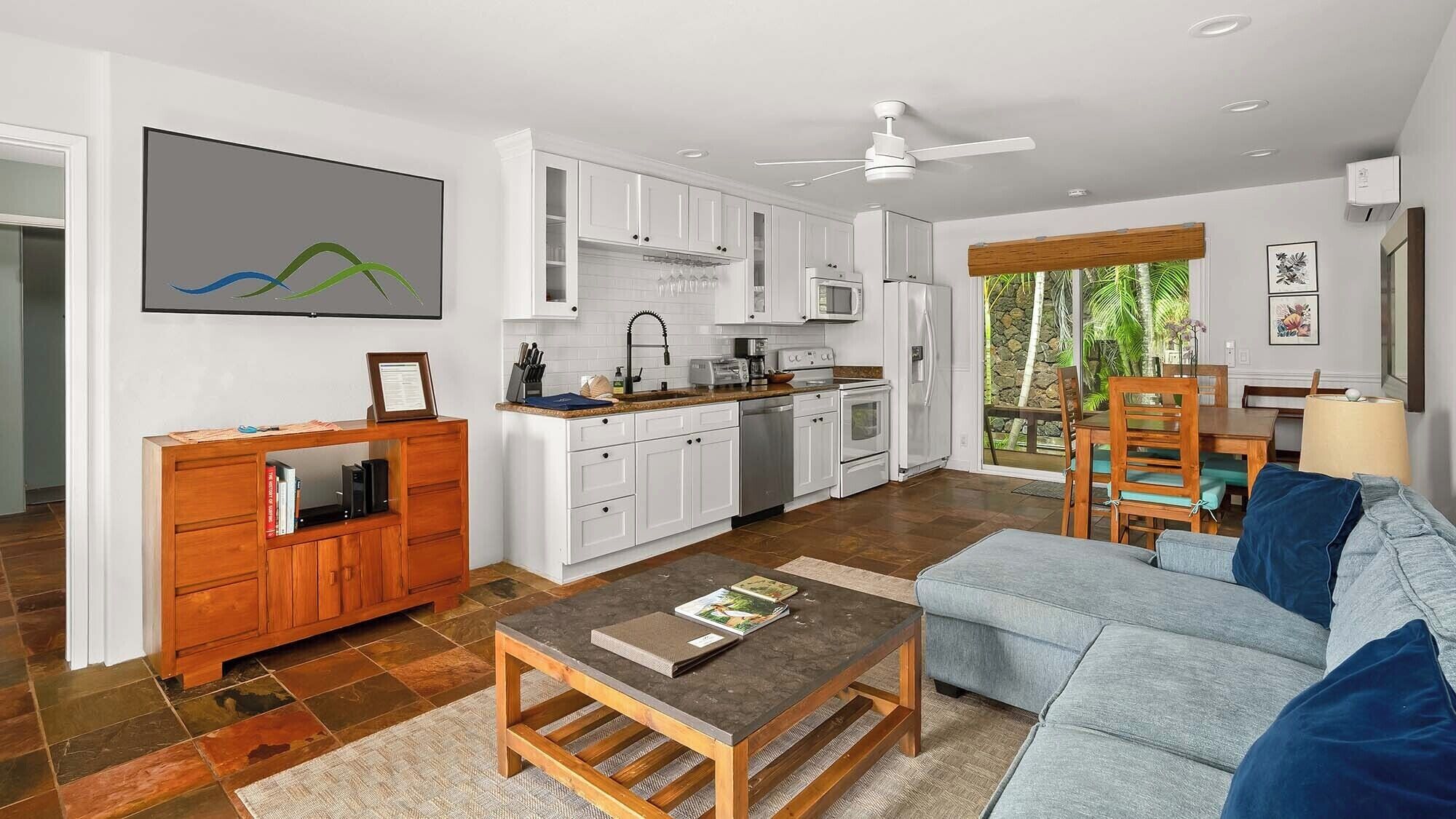 Une cuisine moderne avec des meubles blancs, un coin repas doté d’une table et de chaises en bois, un canapé gris et un ventilateur de plafond.