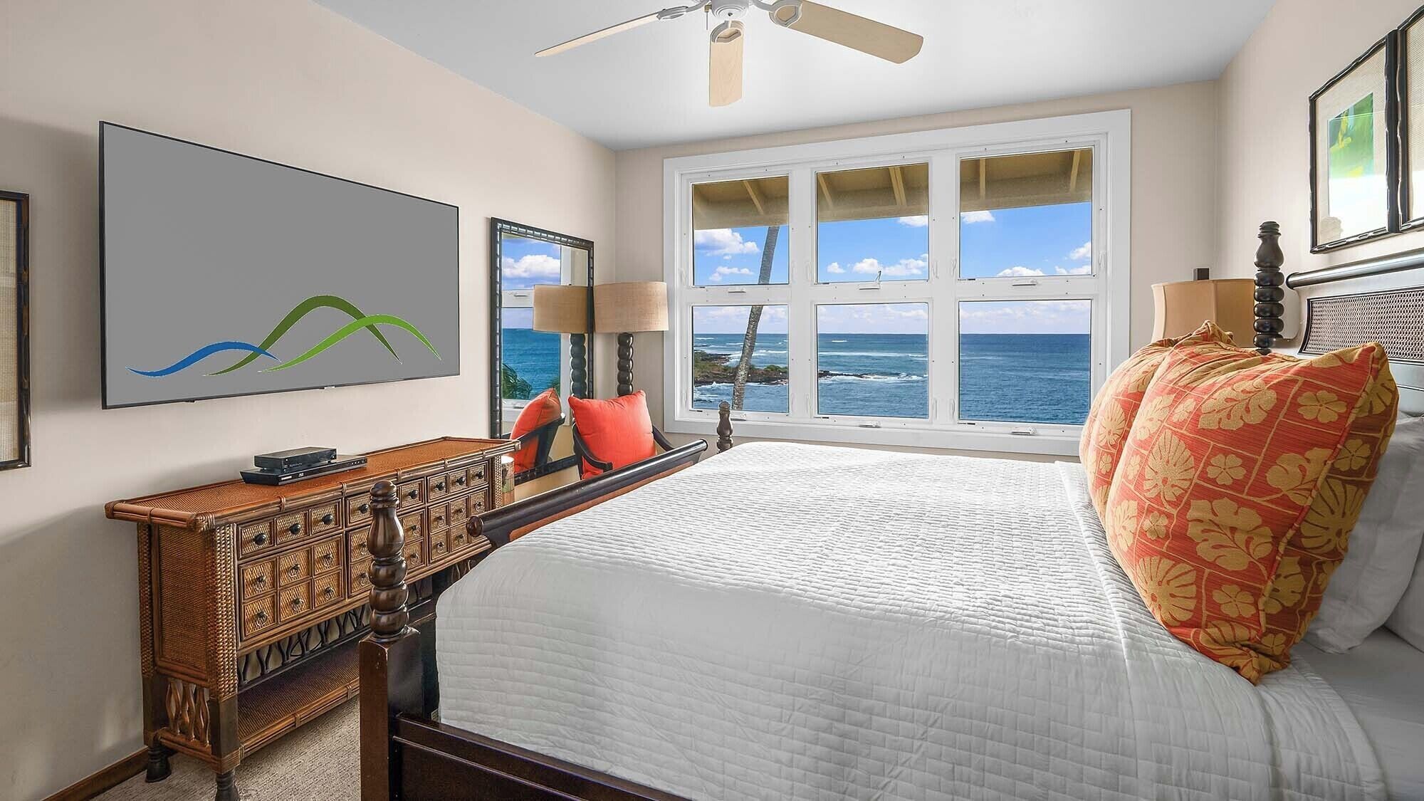 A bedroom with a large bed, a wooden dresser, a ceiling fan, a large window with a view of the ocean, and a framed artwork on the wall.