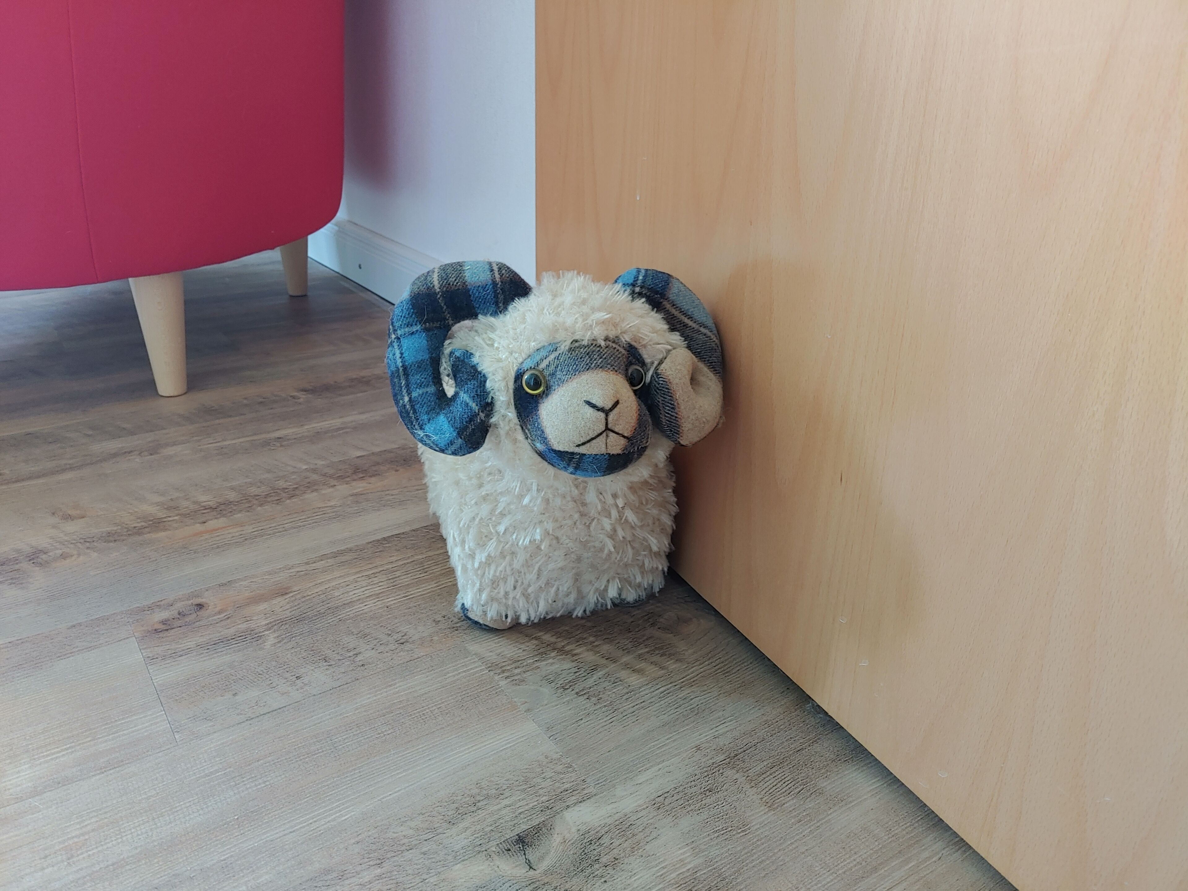 A plush toy sheep with blue and black plaid ears, positioned on a wooden floor next to a wooden wall.