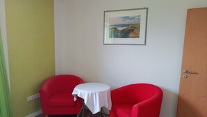 Ein Zimmer mit zwei roten Stühlen, einem kleinen weißen Tisch, einem gerahmten Bild an der Wand und einer Holztür.