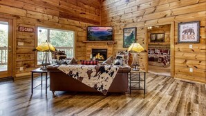 A cabin interior with a fireplace, wooden walls, a couch with a cowhide blanket, and a TV mounted on the wall. - Relaxing Cabin Getaway! HOT TUB! Large Yard! Wifi (Sevierville)