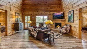 A log cabin interior with a fireplace, a sofa, a television, and a sign reading "CAMPING CABIN." - Relaxing Cabin Getaway! HOT TUB! Large Yard! Wifi (Sevierville)