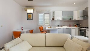 Eine moderne Küche mit weißen Schränken, ein Essbereich mit orangefarbenen Stühlen und ein beiges Sofa.