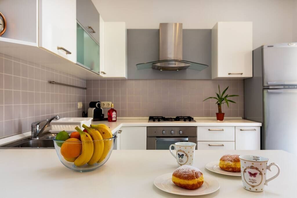 A modern kitchen with white cabinets, a gas stove, and a stainless steel range hood. There is a bowl of fruit and two pastries on the counter.