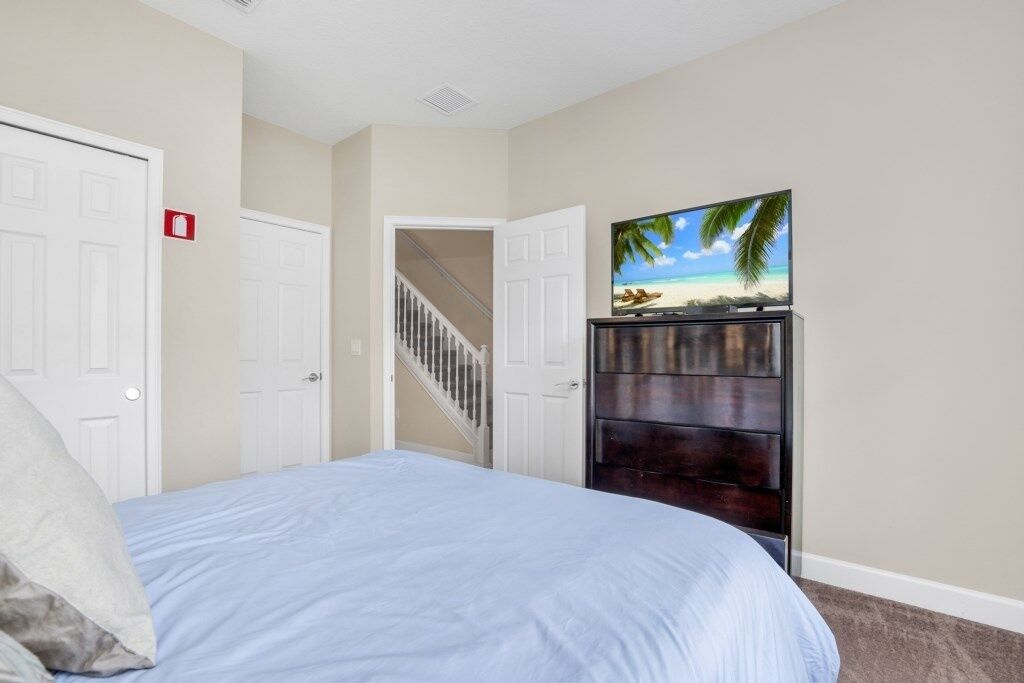 A bedroom with a bed, a TV on a wooden stand, a staircase, and a door with a fire alarm.