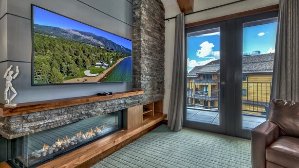 Un salon moderne doté d’un grand téléviseur à écran plat fixé sur un mur en pierre, d’une cheminée avec une façade en verre et offrant une vue sur un paysage montagneux grâce à de grandes portes-fenêtres coulissantes.
