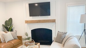 A modern living room with a fireplace, a flat-screen TV mounted above it, a sofa, a coffee table, and a floor lamp. - Charleston "Chucktown Cottage" mins to Beach (Mount Pleasant)