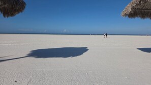 A beach with a clear sky, a few people walking, and a shadow of a person on the sand.